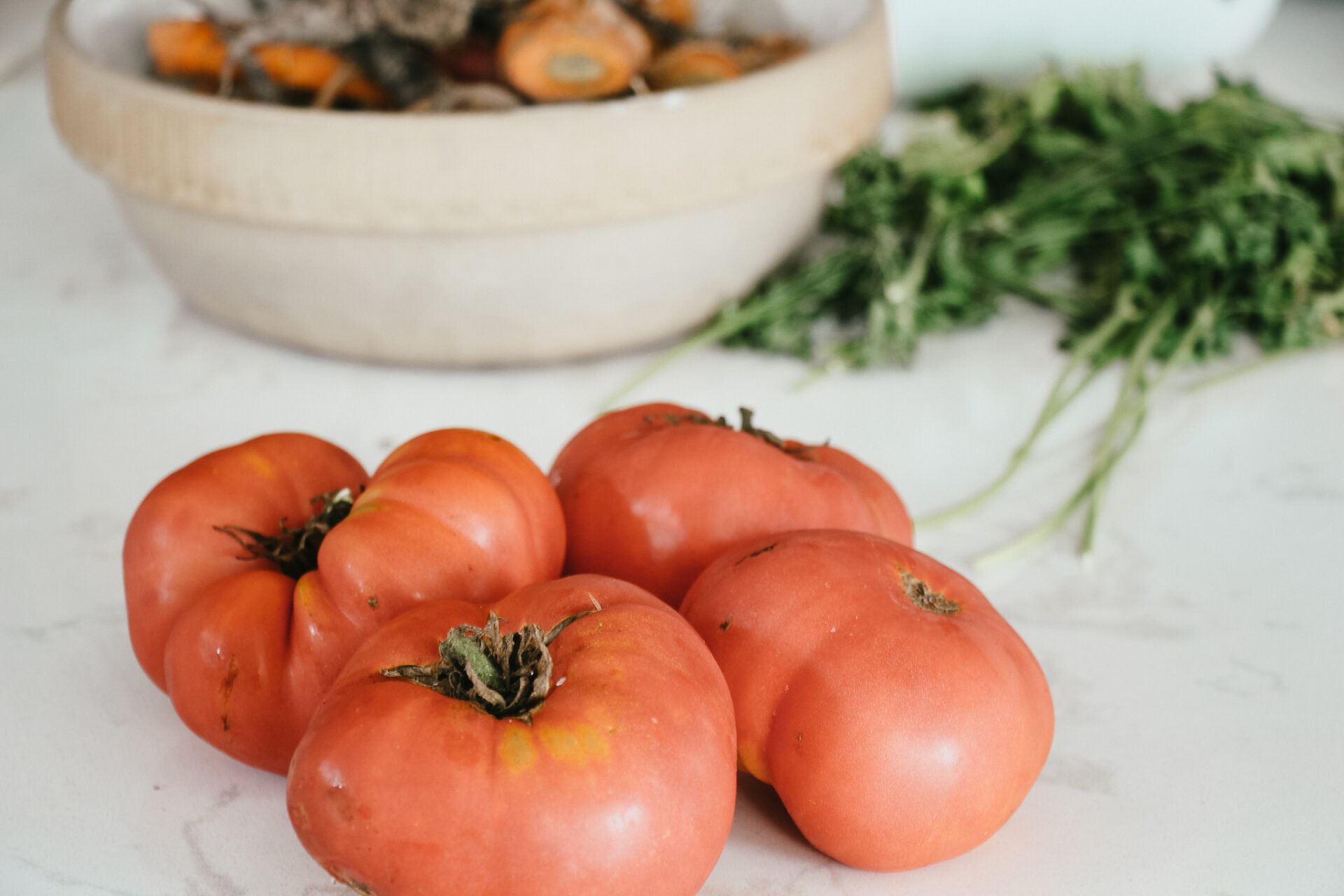 four large red tomatoes sitting on a white marble counter top with other vegetables in the back