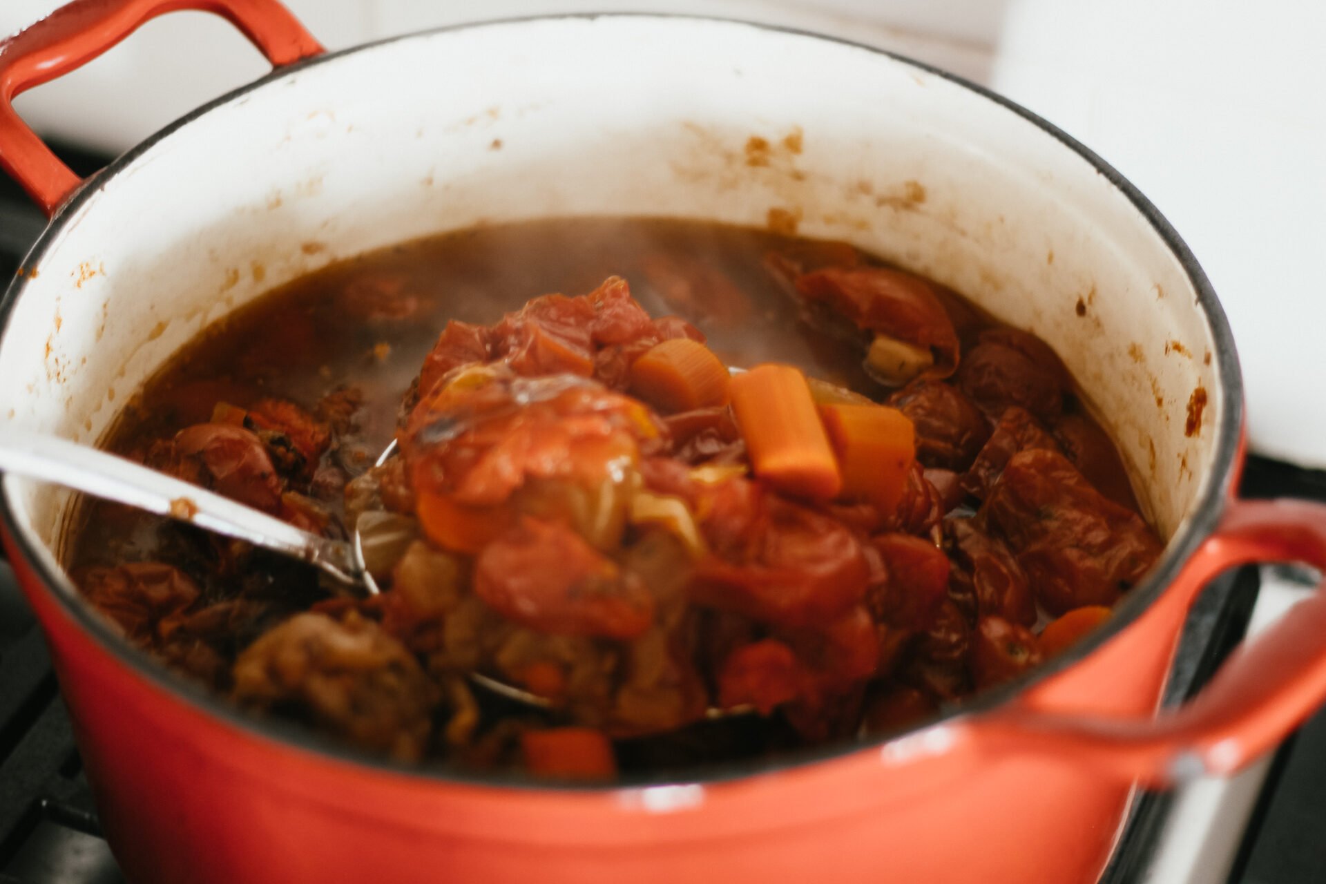 a metal spoon holding a spoonful of marinara sauce from a red dutch oven over the gas stove