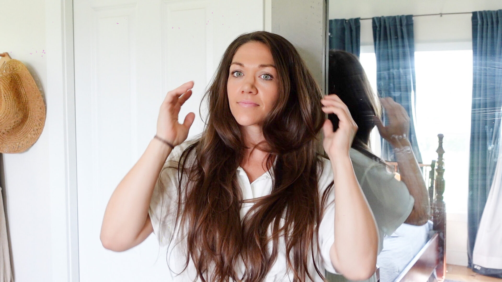 a woman with dark brown hair and a white blouse on looking at her long layered hair with her hands above her ears